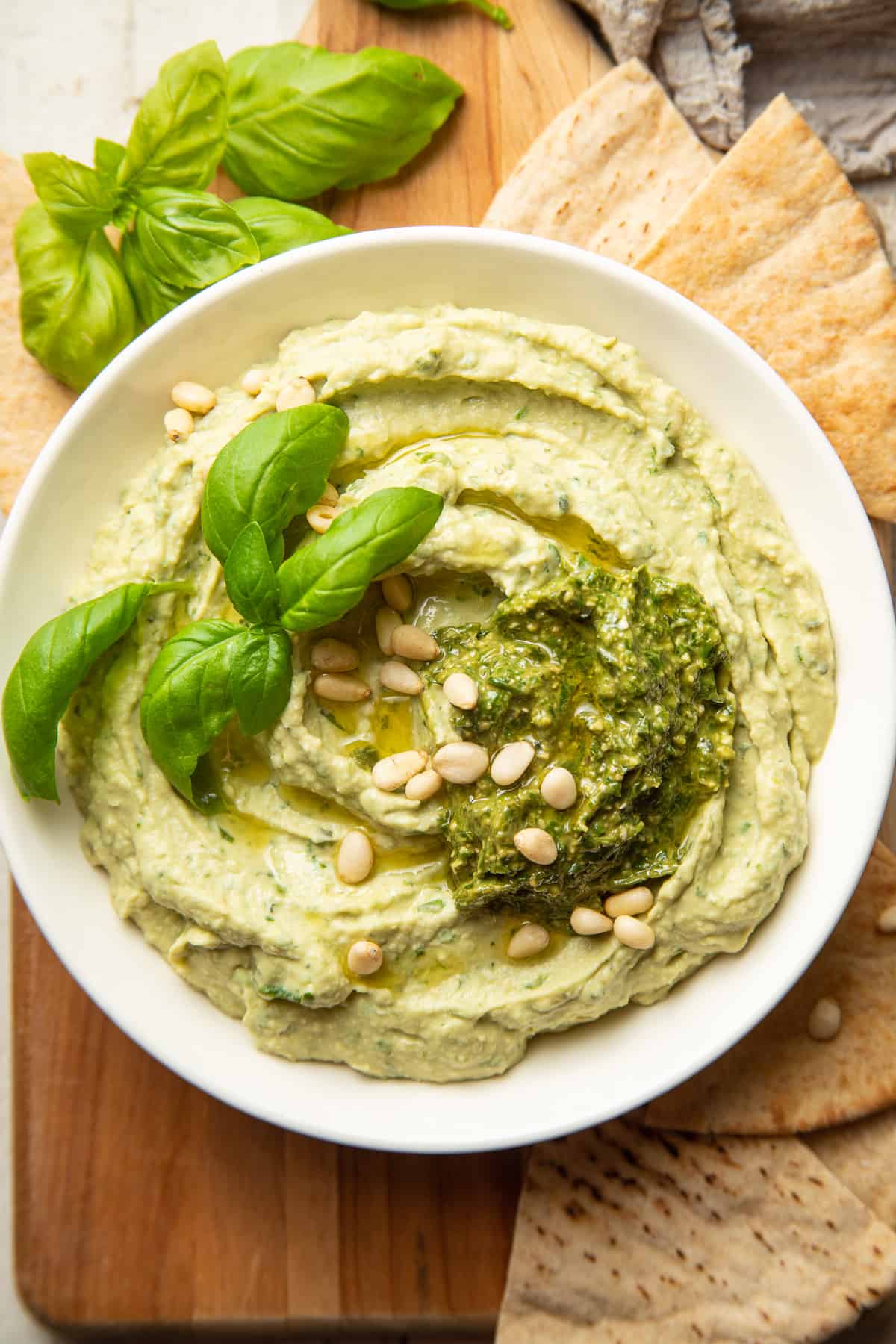 Bowl of Pesto Hummus with pesto, fresh basil and pine nuts on top, and pita wedges on the side.