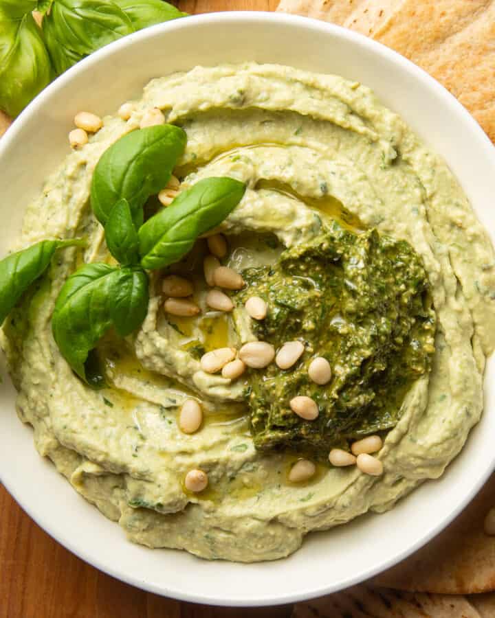 Pesto Hummus in a bowl topped with pine nuts, pesto, and basil leaves.