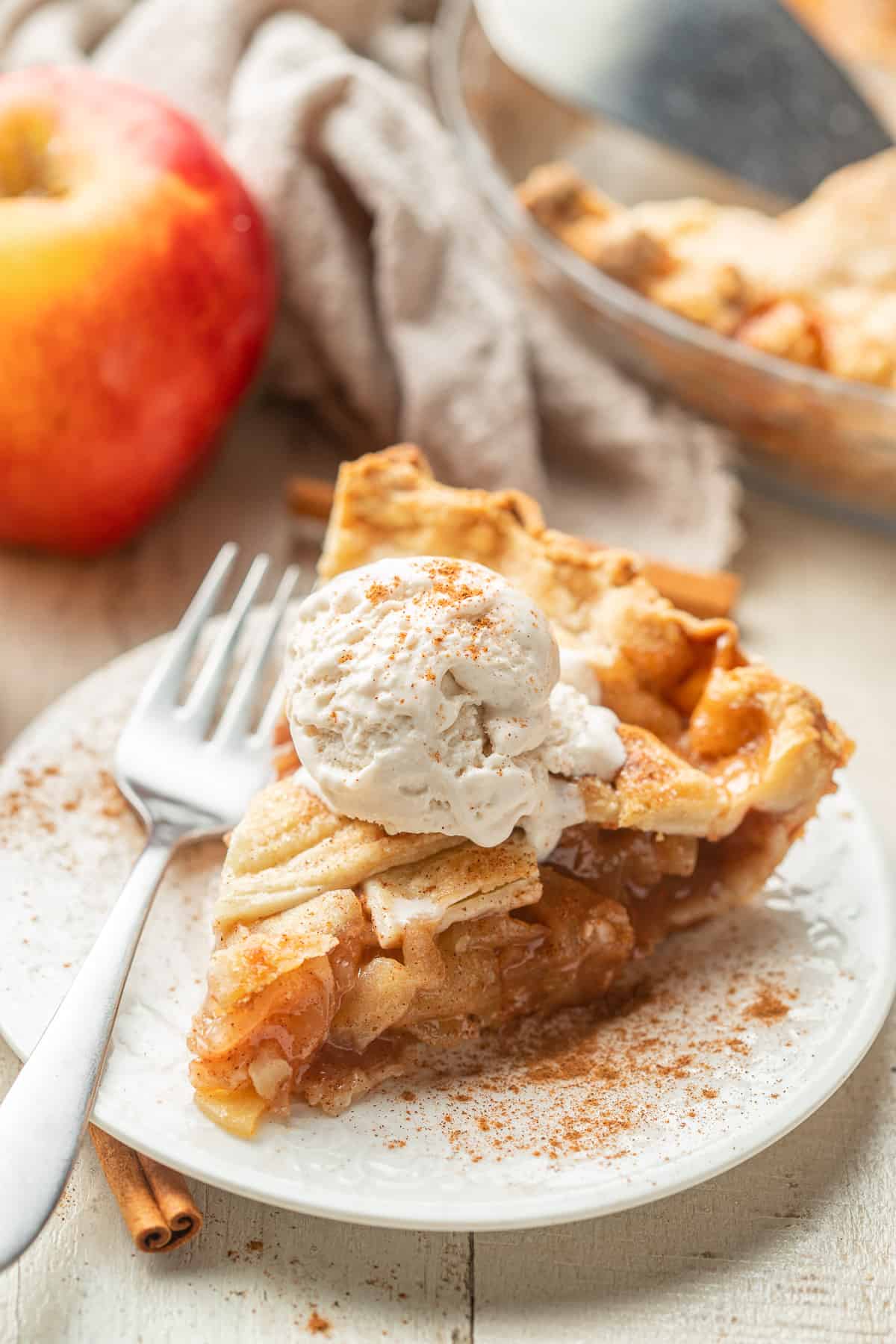 Slice of Vegan Apple Pie with apple and pie dish in the background.