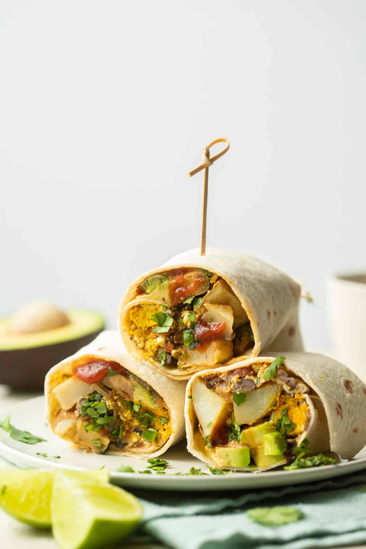 Three Vegan Breakfast Burrito halves stacked on a plate with lime wedges in the foreground.