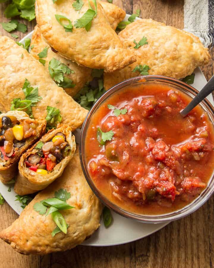 Vegan Empanadas on a plate with salsa, one empanada cut in half to show fillings.