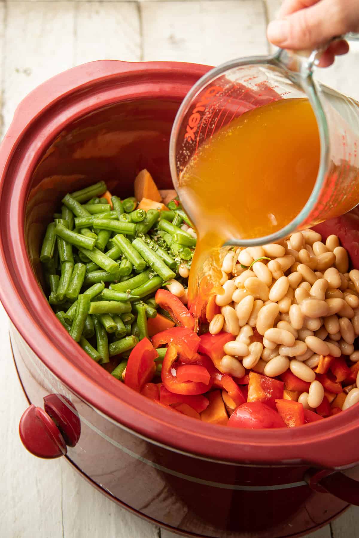 Hand pouring broth into a slow cooker filled with vegetables and beans.