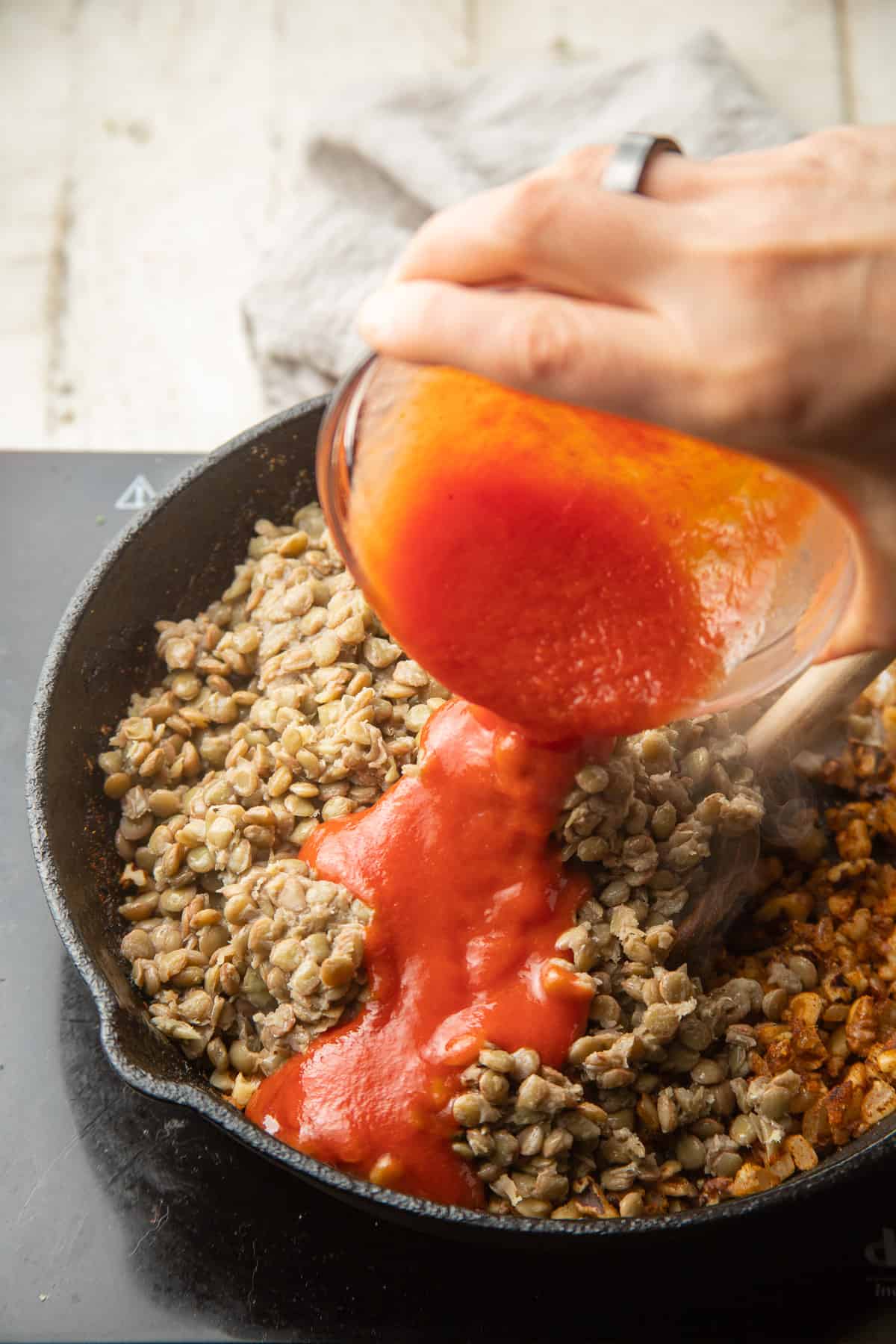 Hand pouring tomato sauce into a skillet of lentil taco filling.