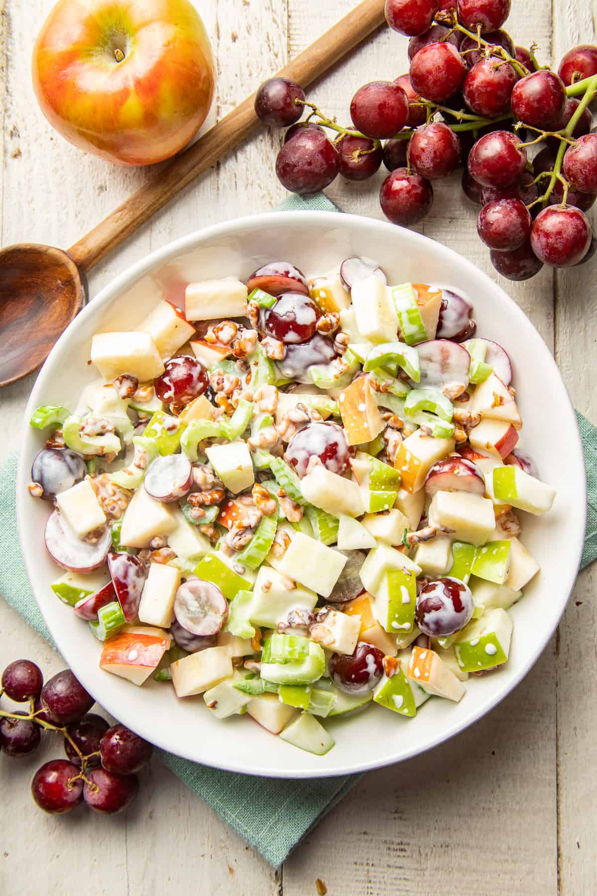 White wooden surface set with grapes, apple, and a bowl of Vegan Waldorf Salad.