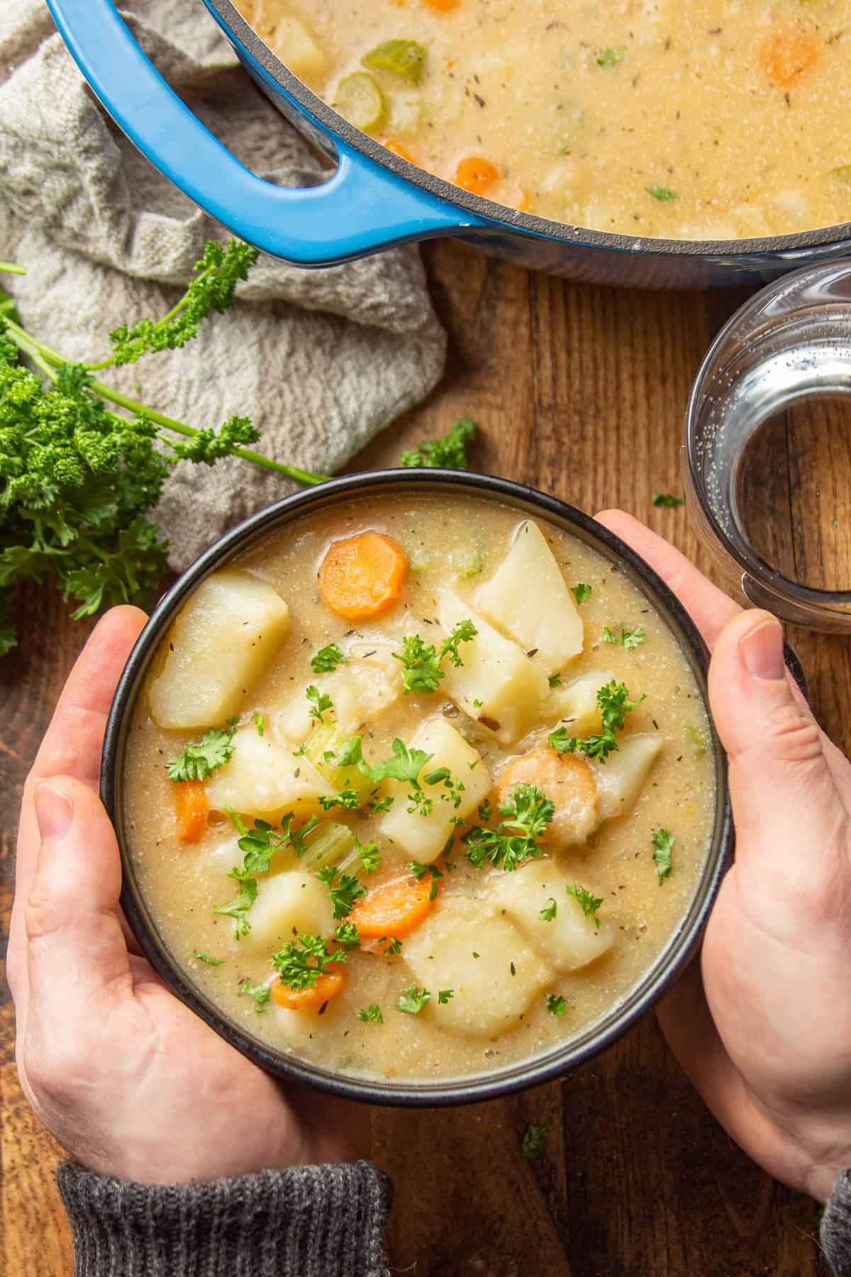Pair of hands holding a bowl of Vegan Potato Soup over a wooden table.