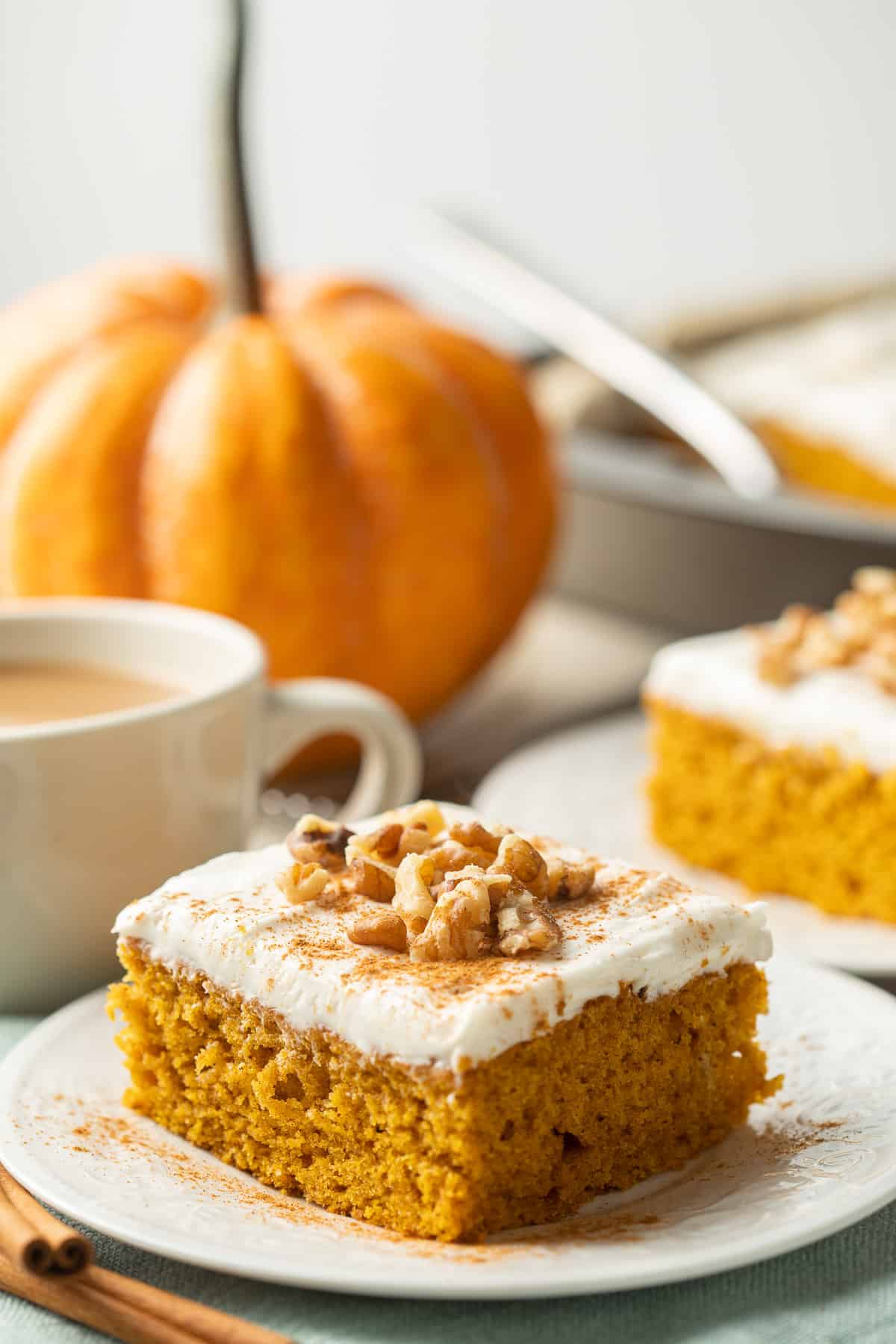 Slice of Vegan Pumpkin Cake on a plate, with pumpkin, coffee cup and more cake in the background.