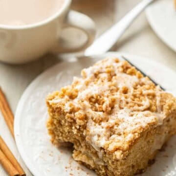 Slice of Vegan Apple Cake on a dish with coffee cup and cinnamon sticks on the side.