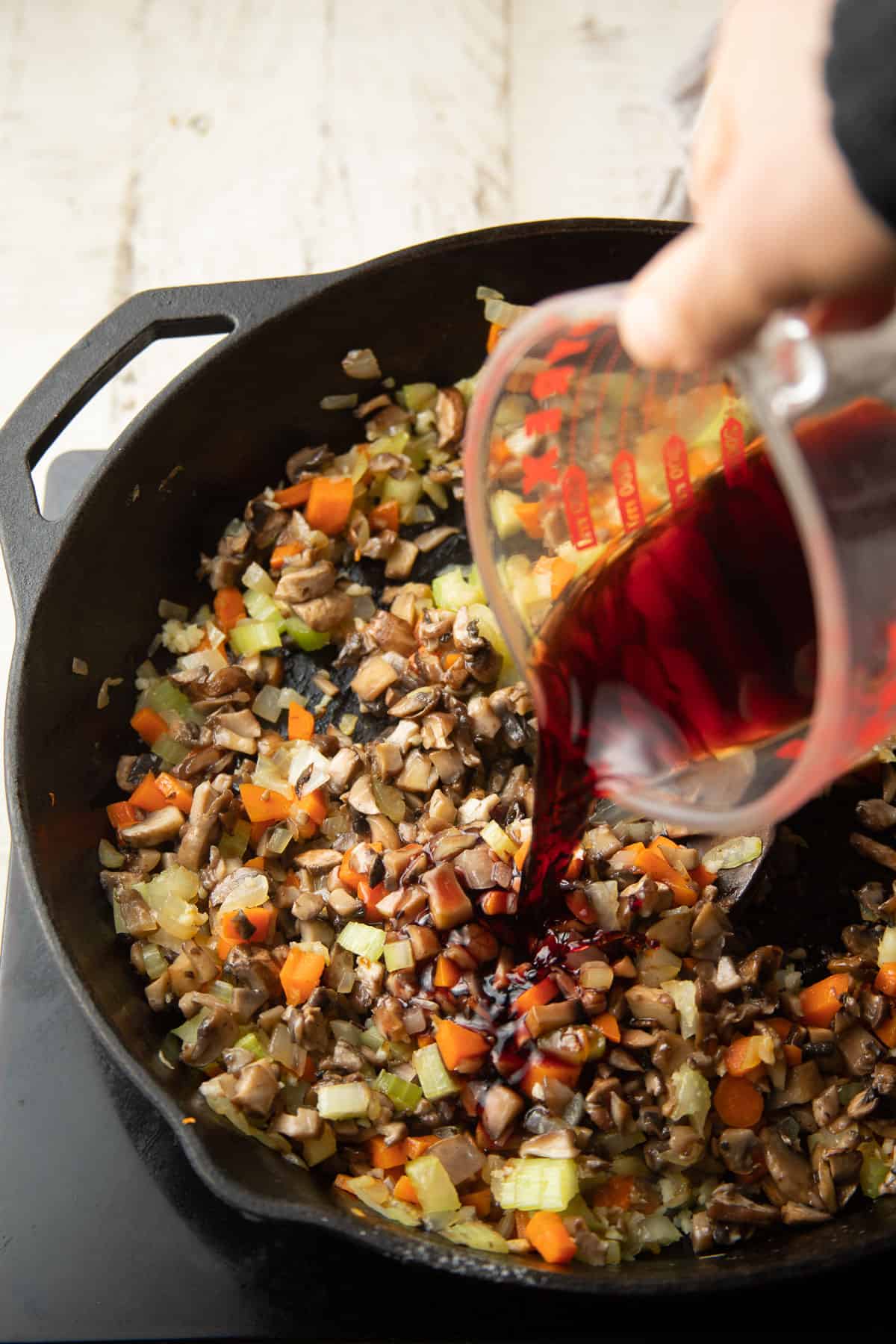 Hand pouring wine into a skillet filled with mushrooms, carrots, onions, and celery.