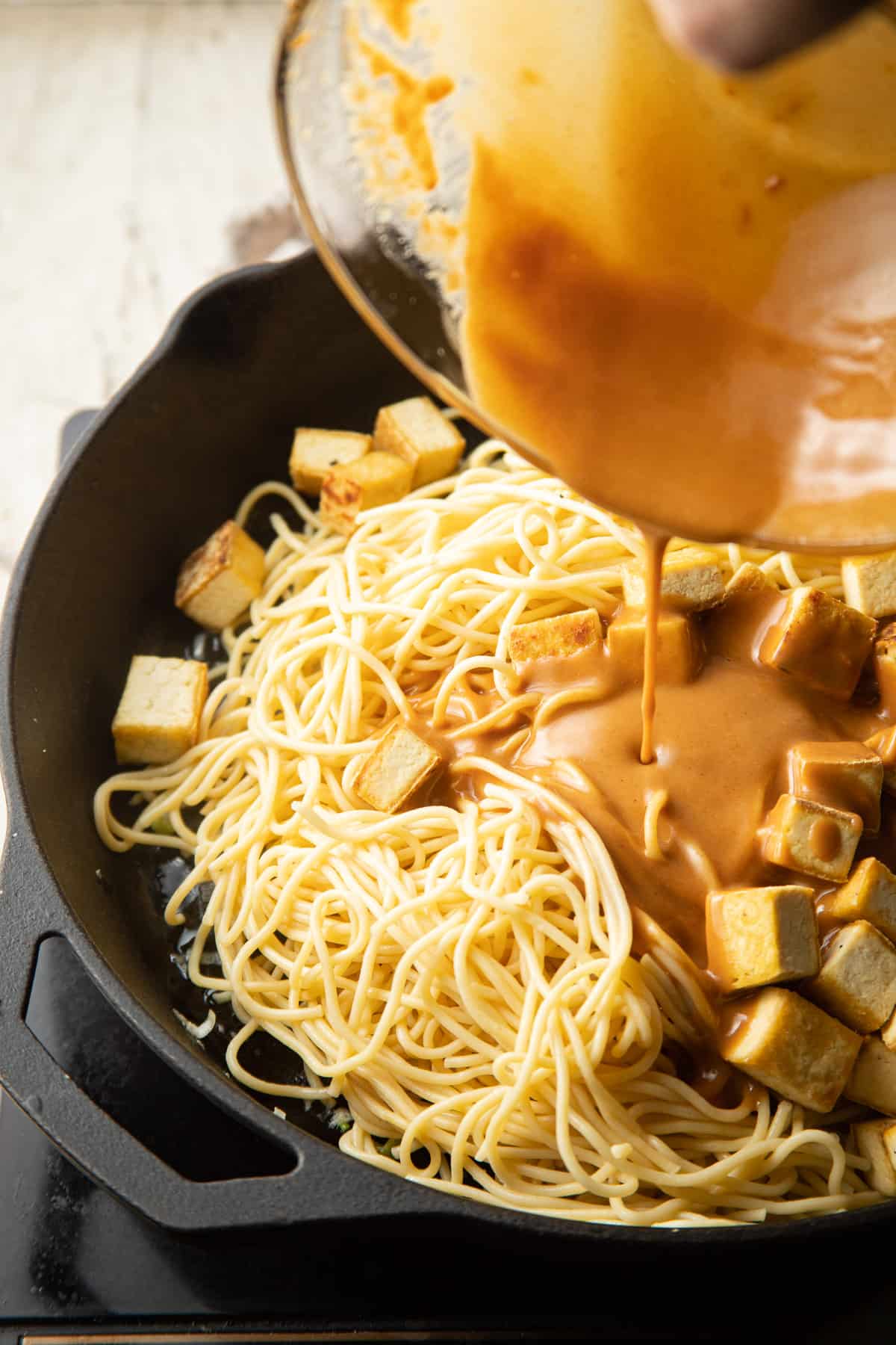 Peanut sauce being poured over noodles and tofu in a skillet.