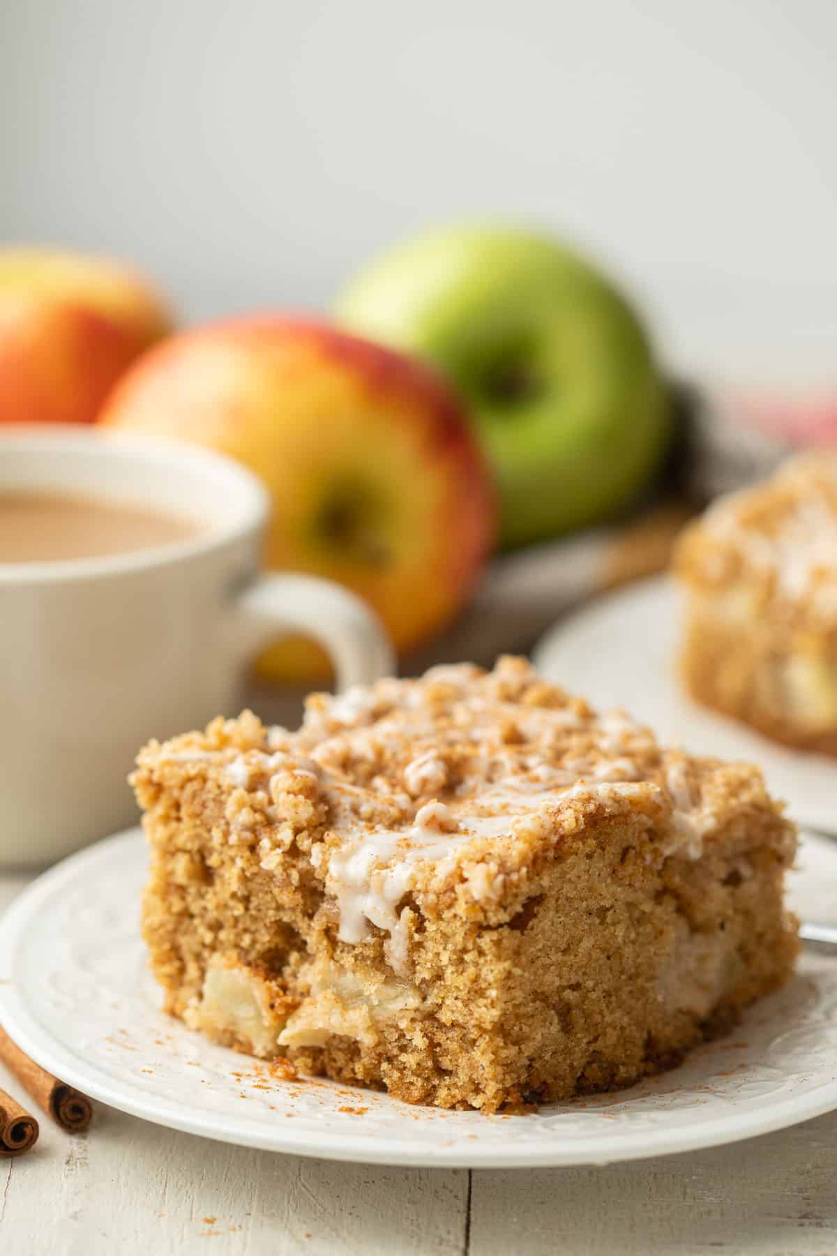 Close up of a slice of Vegan Apple Cake with apples and coffee cup in the background.