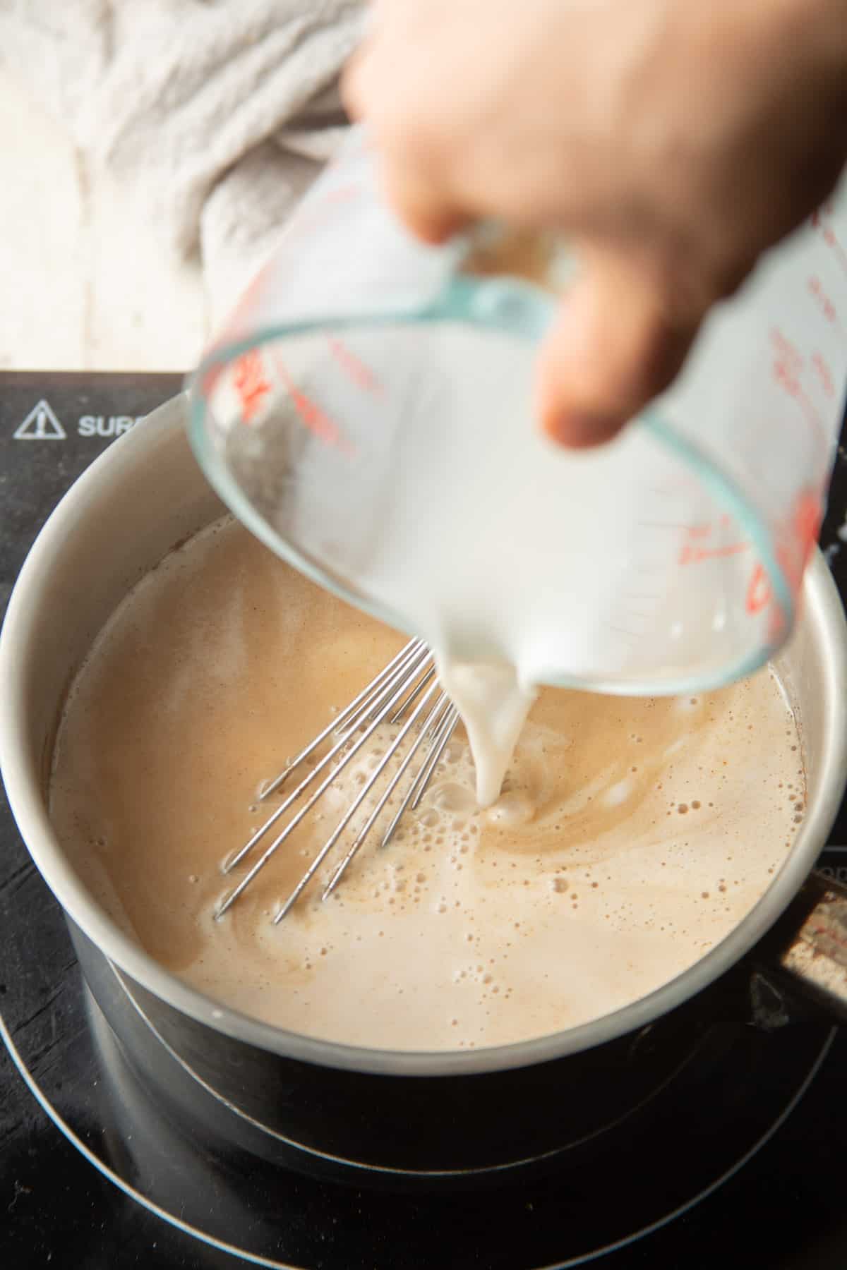 Hand pouring cornstarch slurry into a saucepan of milk and maple syrup.