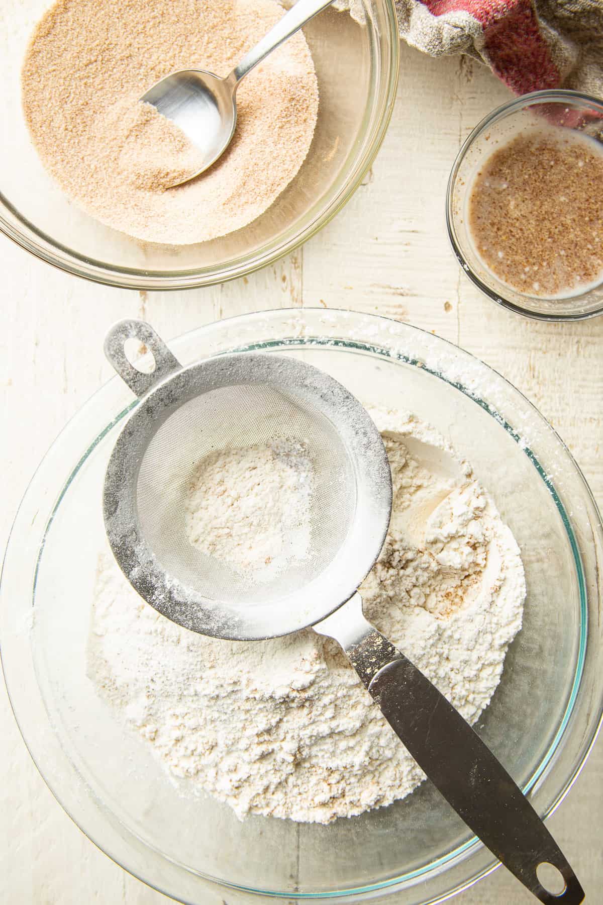 Three glass bowls on a table containing sifted flour, flax egg and cinnamon sugar.
