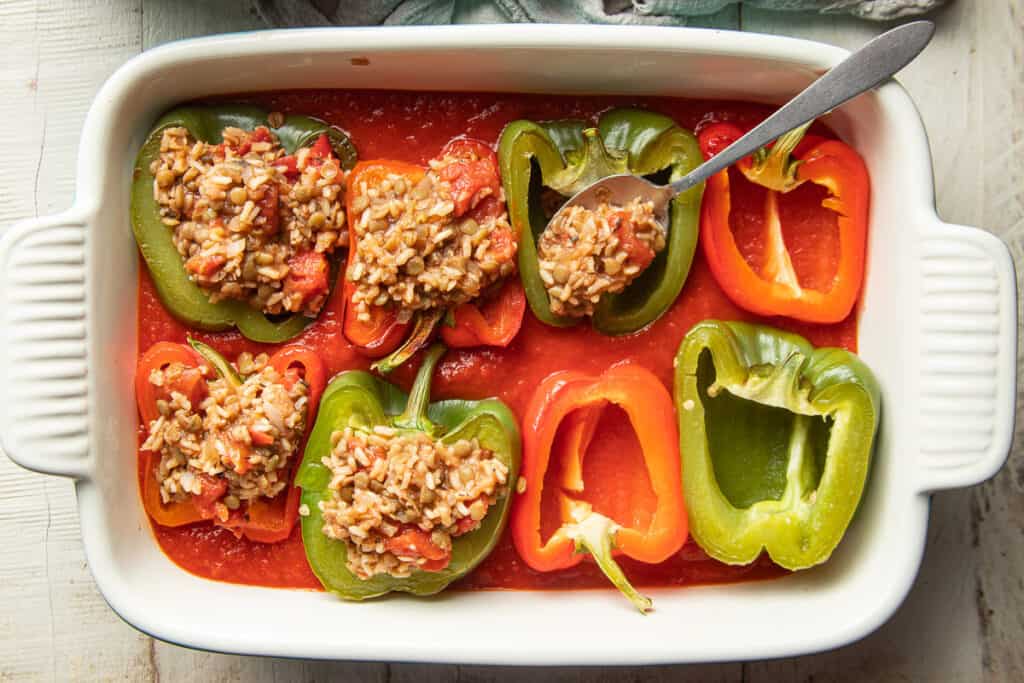 Partially filled bell pepper halves in a baking dish filled with tomato sauce.