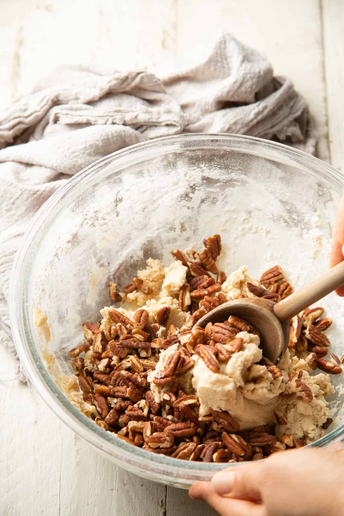 Hand stirring pecans into cookie dough in a glass bowl.