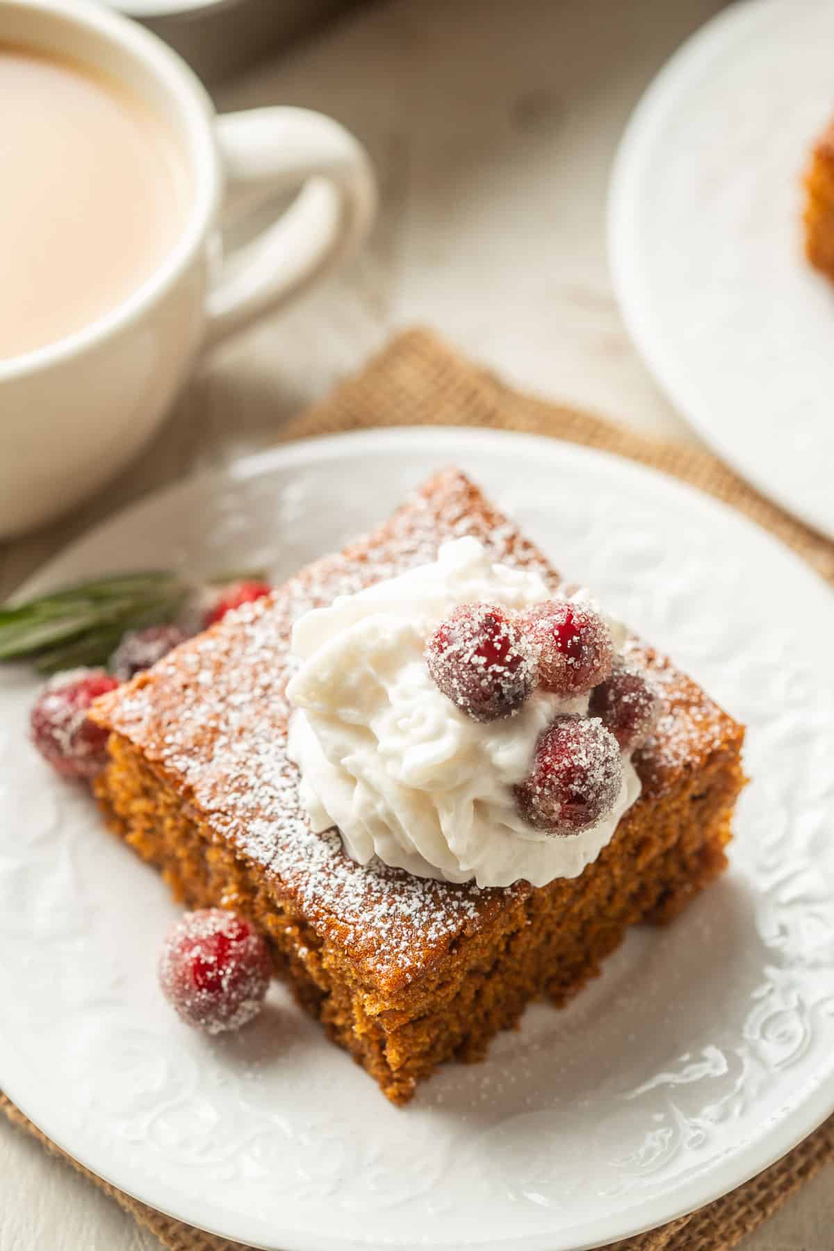 Slice of Vegan Gingerbread Cake on a dish with whipped cream, rosemary and cranberries, with coffee cup in the background.