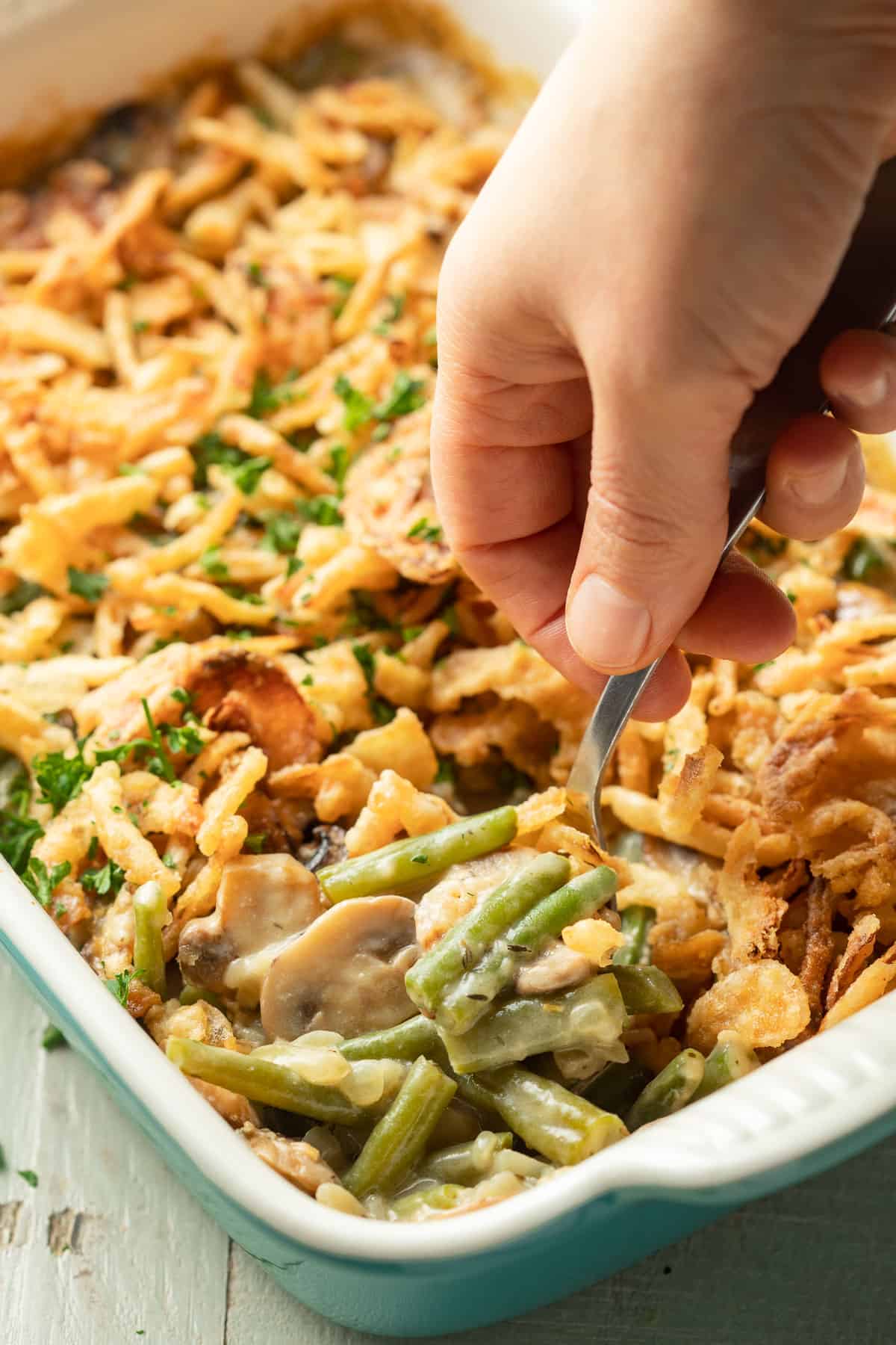 Hand with spoon scooping Vegan Green Bean Casserole from a serving dish.