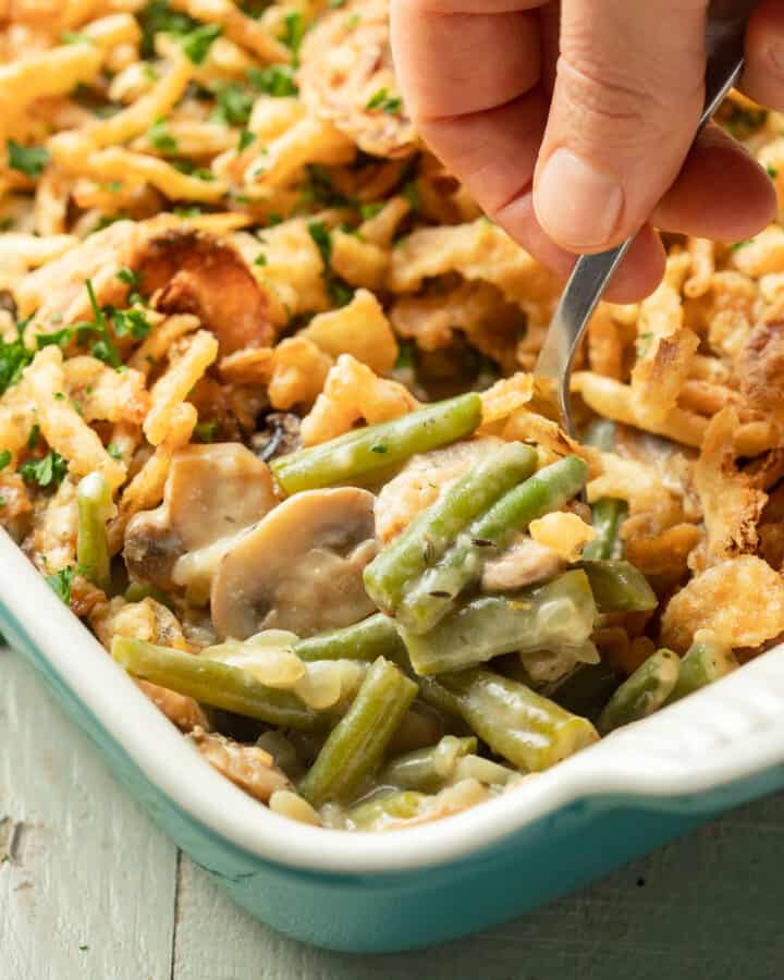 Hand with spoon scooping Vegan Green Bean Casserole from a baking dish.