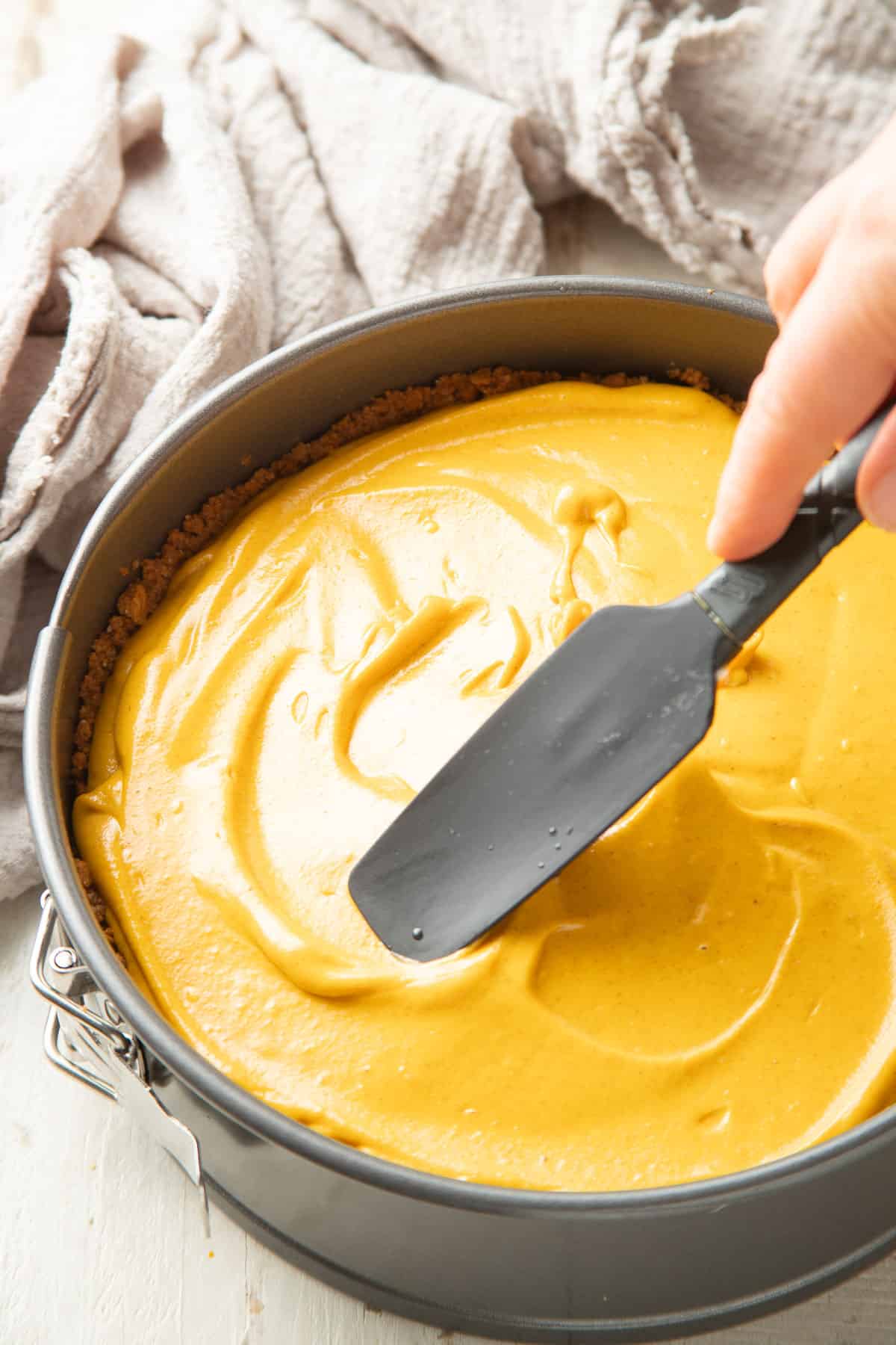 Hand with spatula smoothing Vegan Pumpkin Cheesecake batter in a pan.