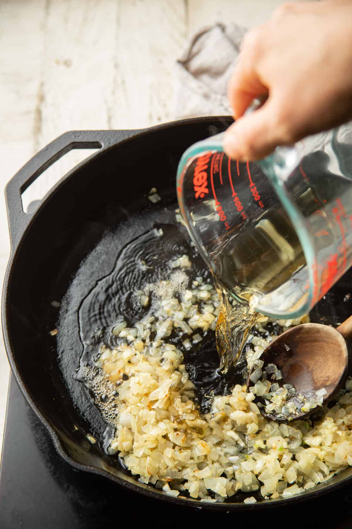 Hand pouring whiskey into a skillet of onions and garlic.