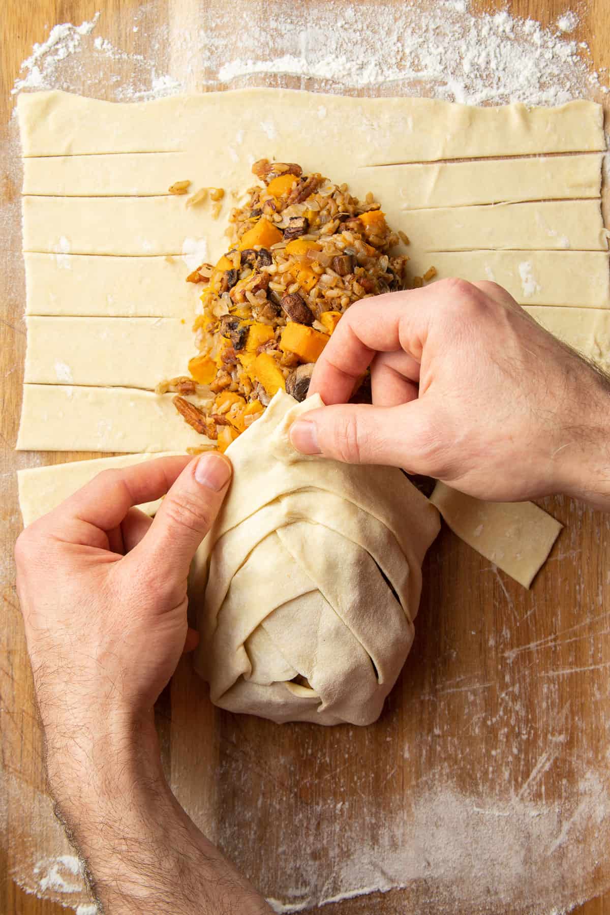Hands braiding puff pastry over Vegetable Wellington filling.