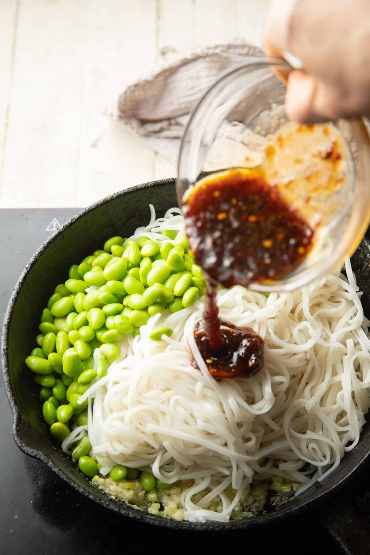 Hand pouring chili garlic sauce into a skillet filled with noodles and edamame.
