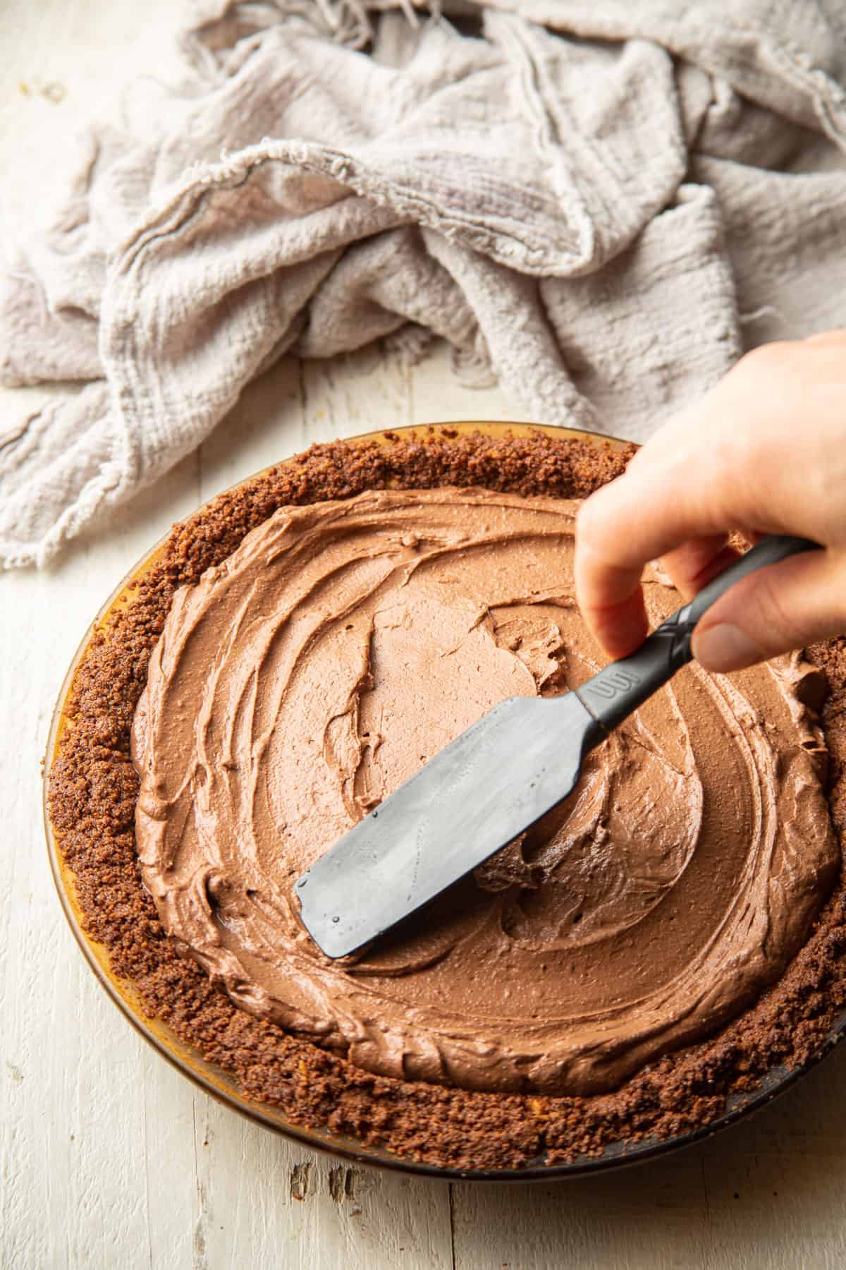 Hand using a spatula to spread chocolate filling in a pie crust.