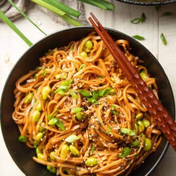 White wooden surface set with skillet, bunch of scallions and bowl of Chili Garlic Noodles.
