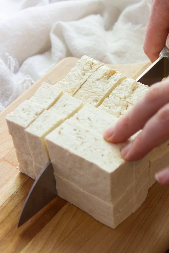 Hand cutting tofu into cubes on a cutting board.