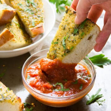 Hand dipping a slice of garlic bread into a bowl of tomato sauce.