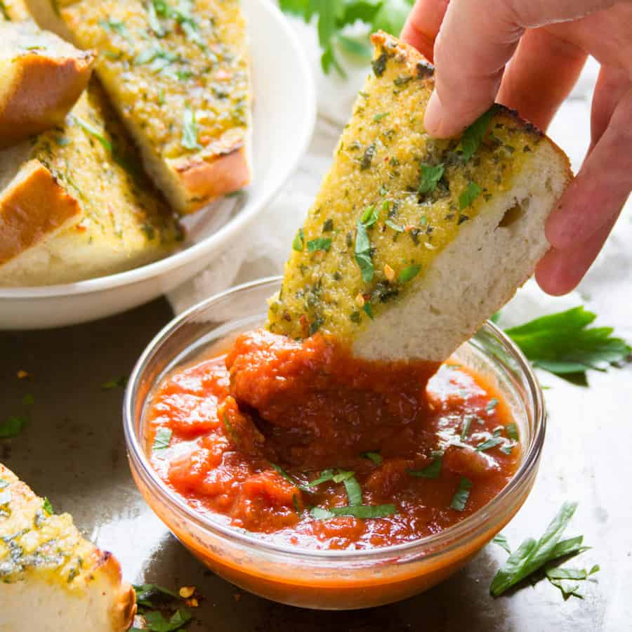 Hand dipping a slice of garlic bread into a bowl of tomato sauce.