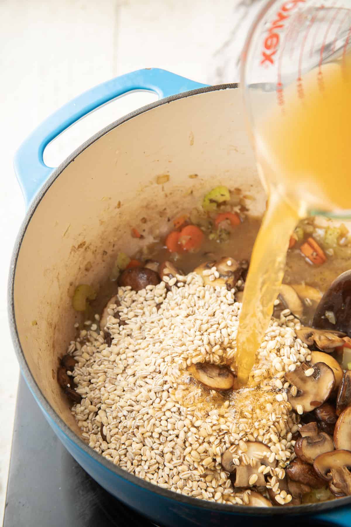 Broth being poured into a pot filled with vegetables, mushrooms, and pearl barley.