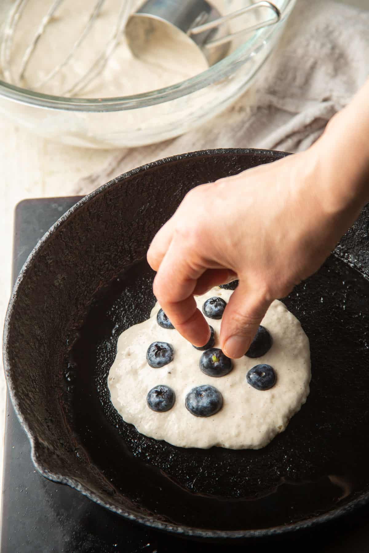 Hand arranging blueberries over a dollop of pancake batter cooking in a skillet.