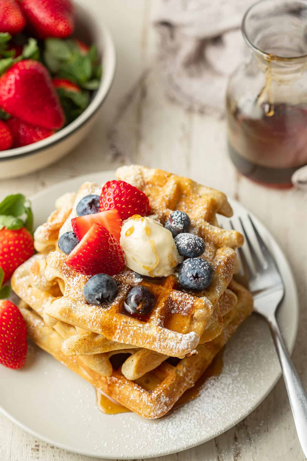 Stack of Vegan Waffles on a plate with bowl of strawberries and container of syrup in the background.