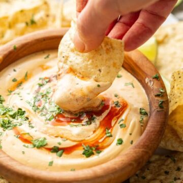 Hand dipping a chip into a bowl of vegan queso topped with hot sauce and cilantro.