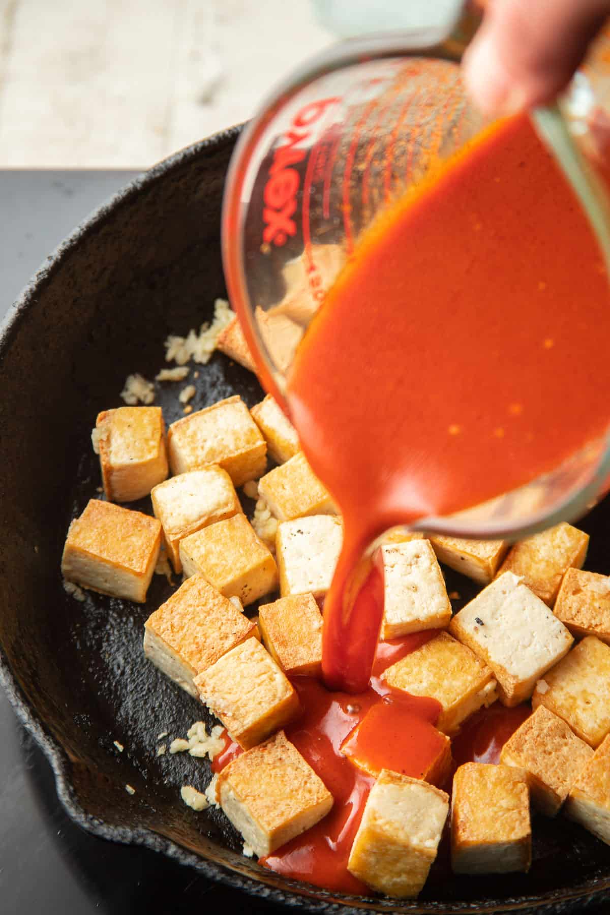 Hand pouring gochujang sauce into a skillet of tofu.