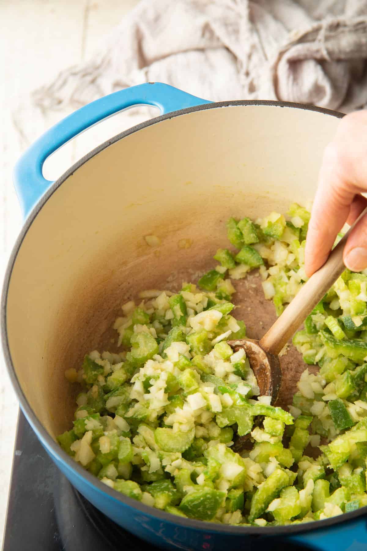 Hand stirring a pot of diced onions, peppers, and celery cooking in a roux.