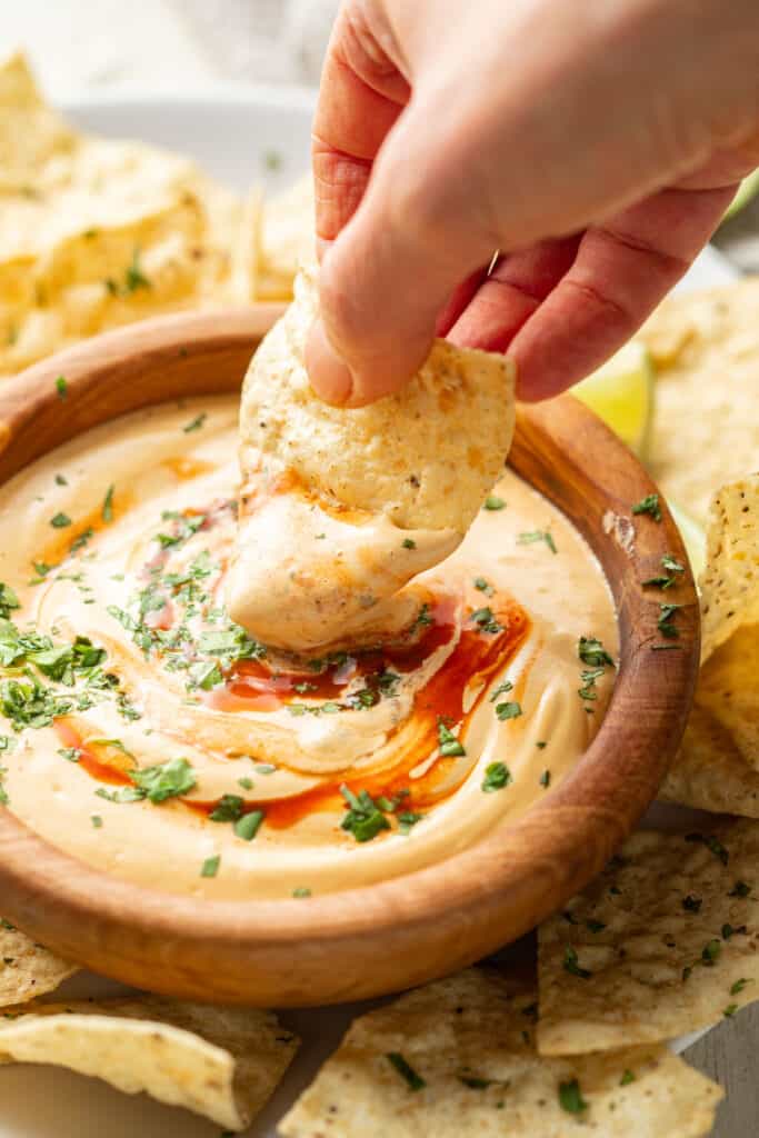 Hand dipping a chip into a bowl of vegan queso topped with hot sauce and cilantro.