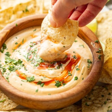 Hand dipping a chip into a bowl of Vegan Cashew Queso topped with hot sauce and cilantro.