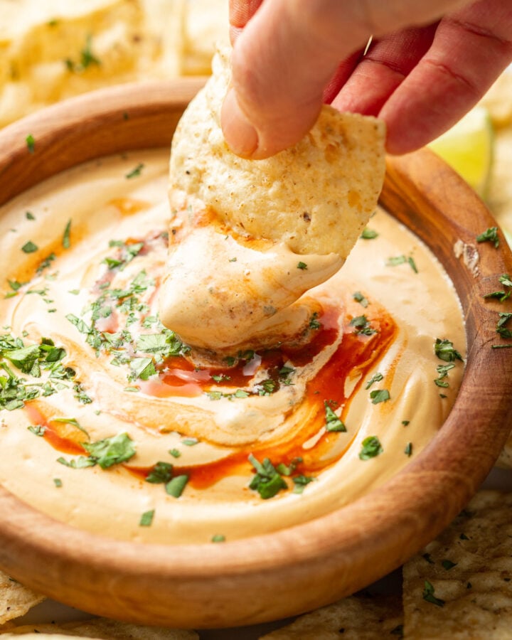 Hand dipping a chip into a bowl of Vegan Cashew Queso topped with hot sauce and cilantro.