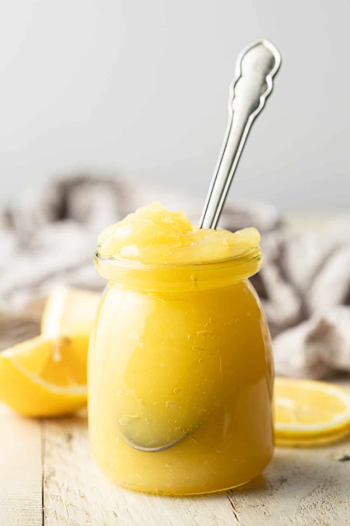 Jar of Vegan Lemon Curd with spoon sticking out, lemons and napkin in the background.