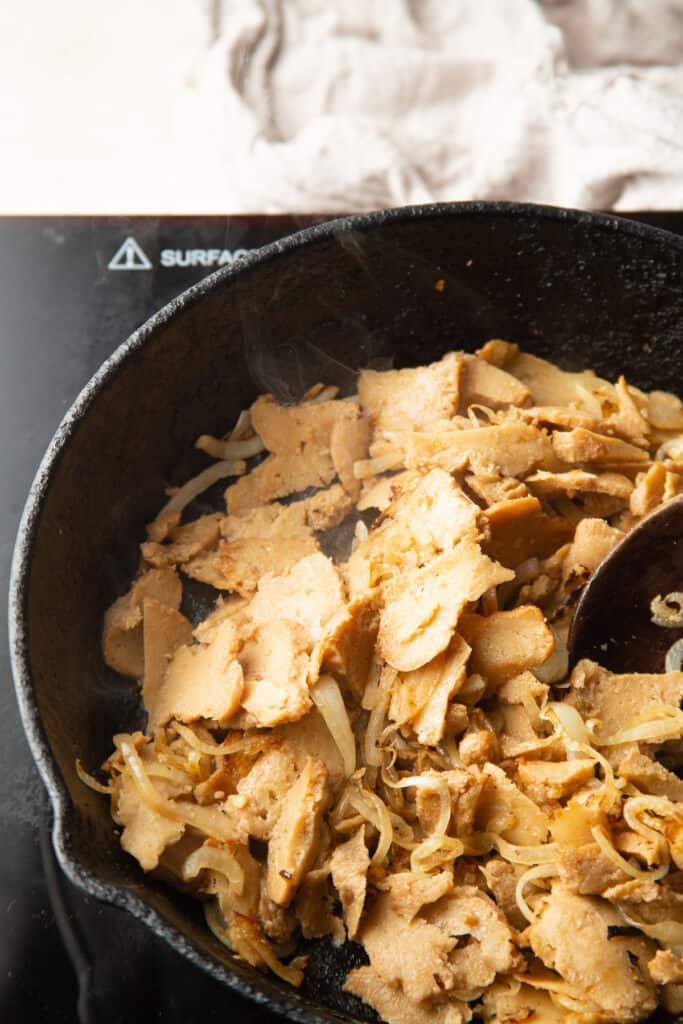 Seitan slices and onions cooking in a skillet.