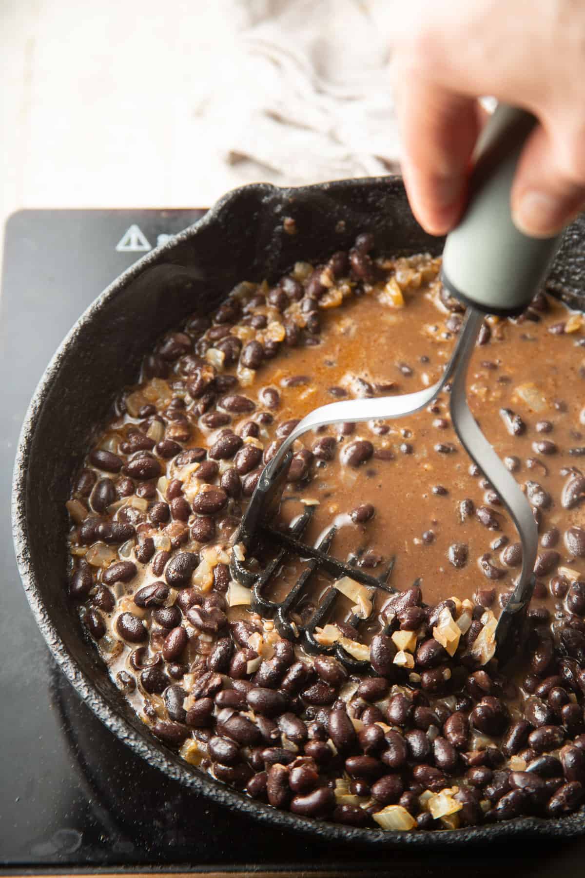 Hand with potato masher mashing beans in a skillet.