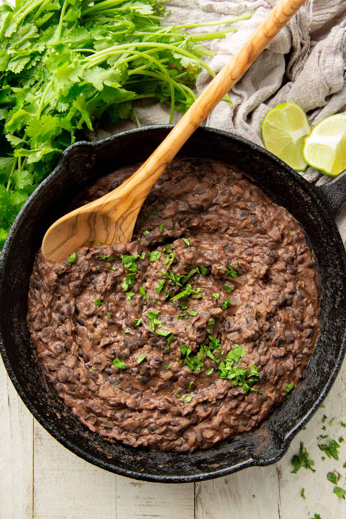 White wooden surface set with skillet of Refried Black Beans, lime slices and bunch of cilantro.