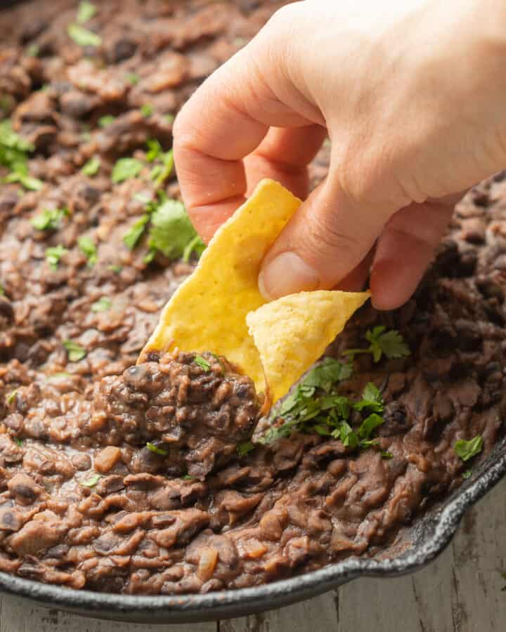 Hand dipping tortilla chip in a skillet of refried black beans.