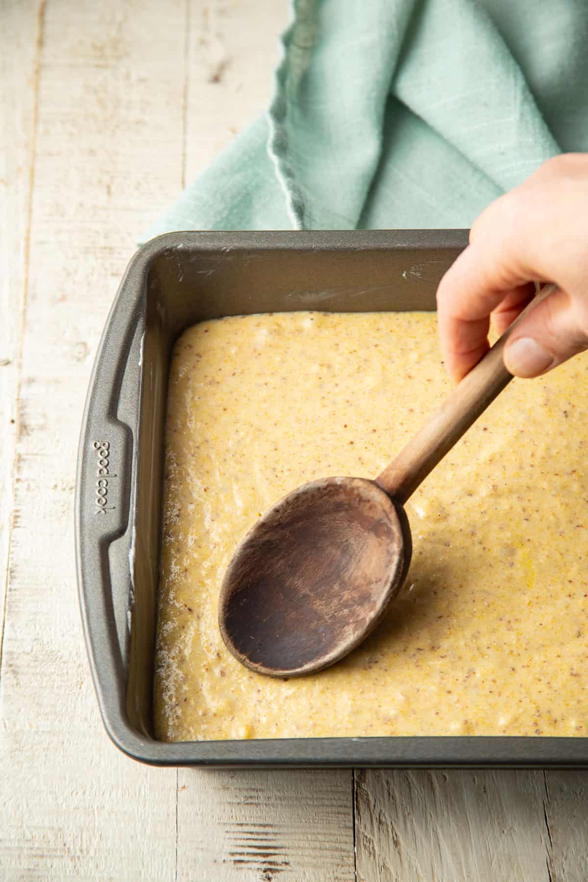 Hand using a wooden spoon to smooth Vegan Cornbread batter in a baking pan.