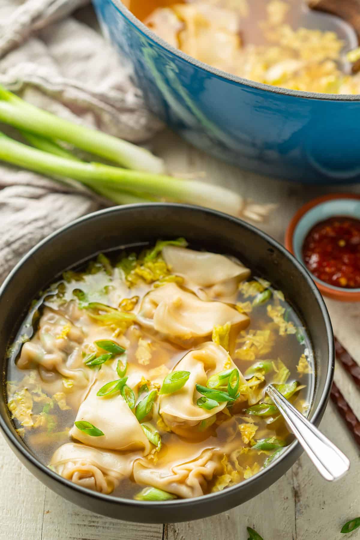 Bowl of Vegan Wonton Soup with blue pot and bunch of scallions in the background.