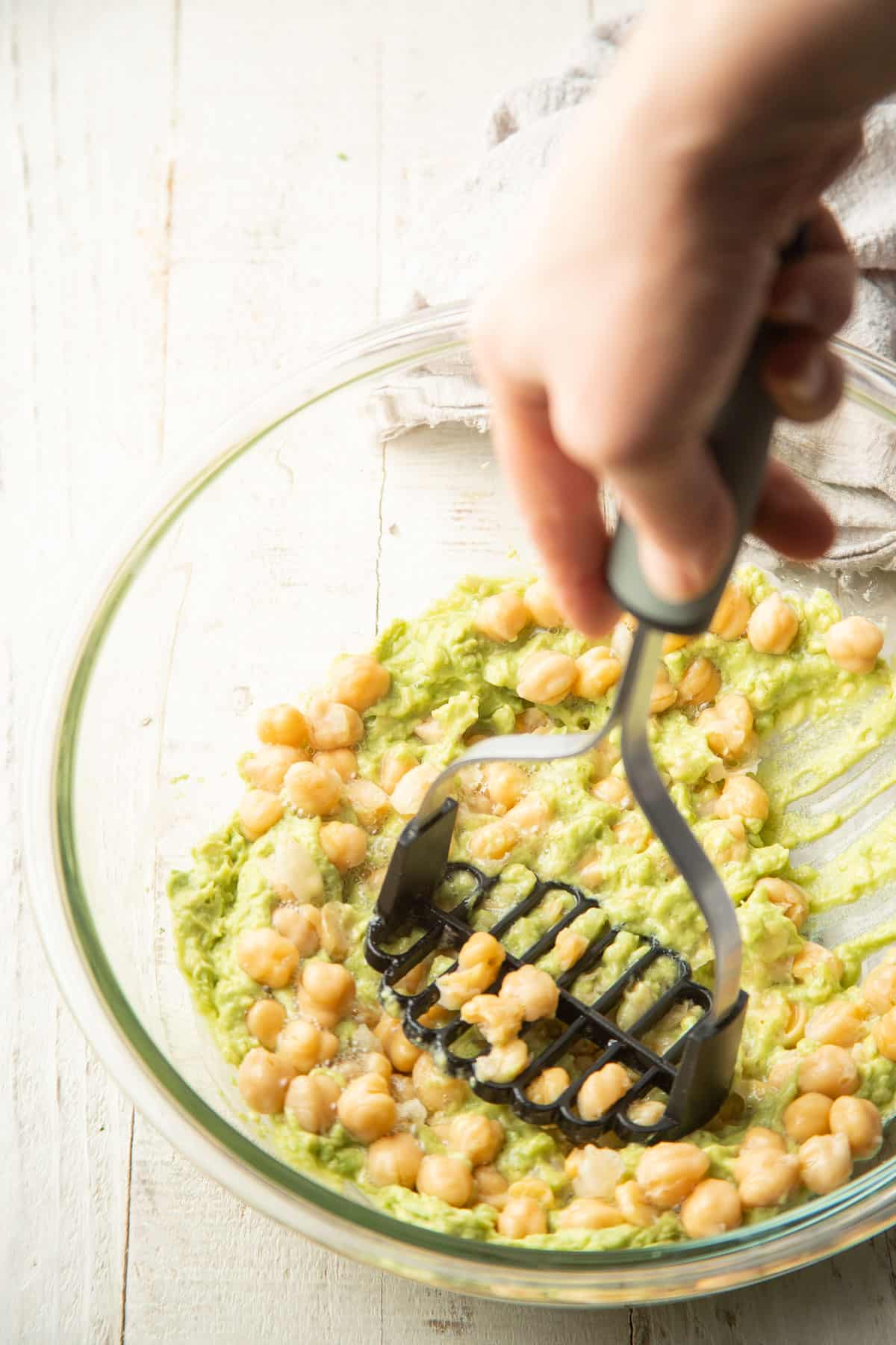 Hand with a potato masher smashing chickpeas in a bowl with avocado dressing.