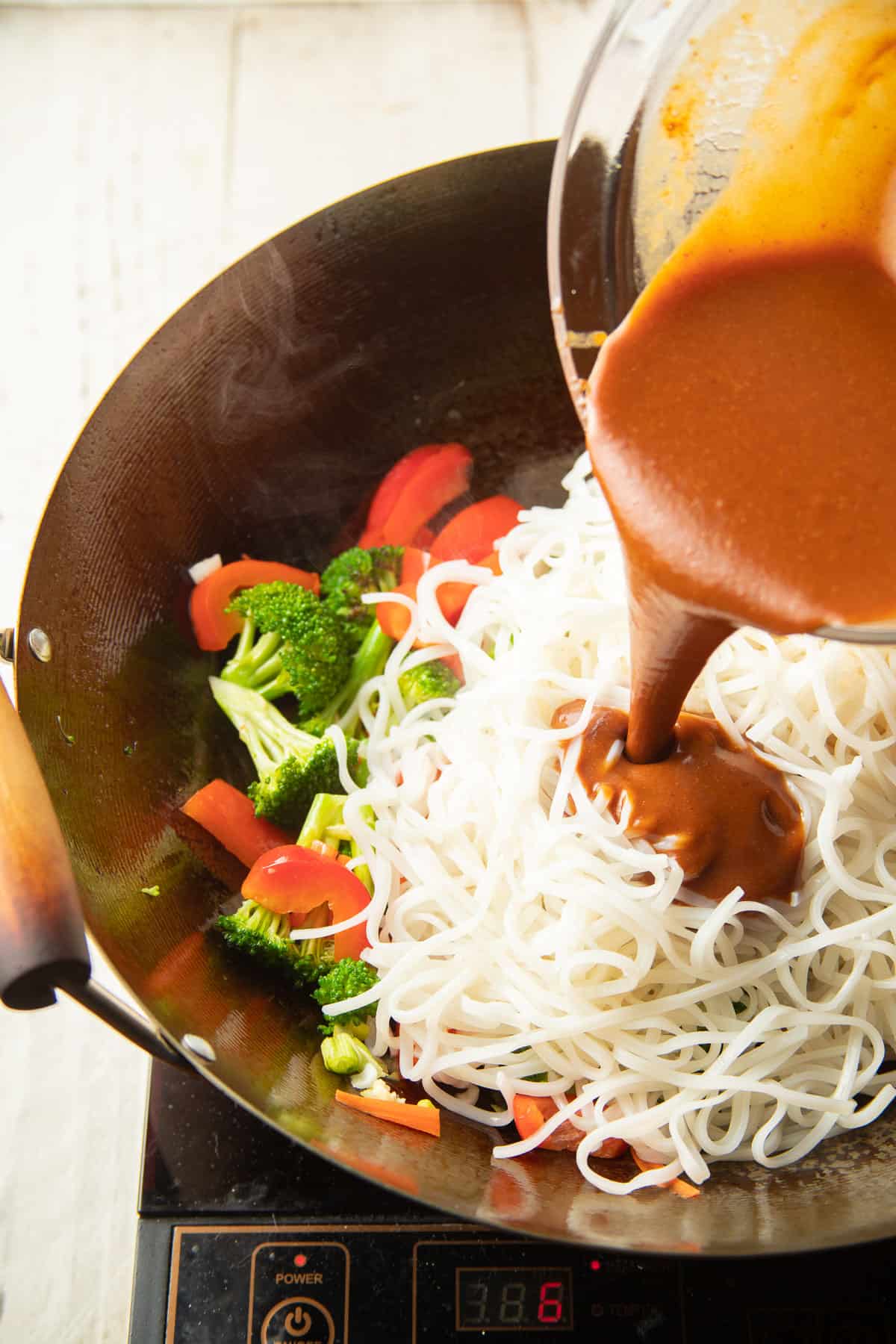 Sauce being poured into a wok filled with vegetables and noodles.