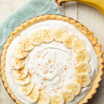 White wooden surface set with bunch of bananas, blue napkin and a whole Vegan Banana Cream Pie.