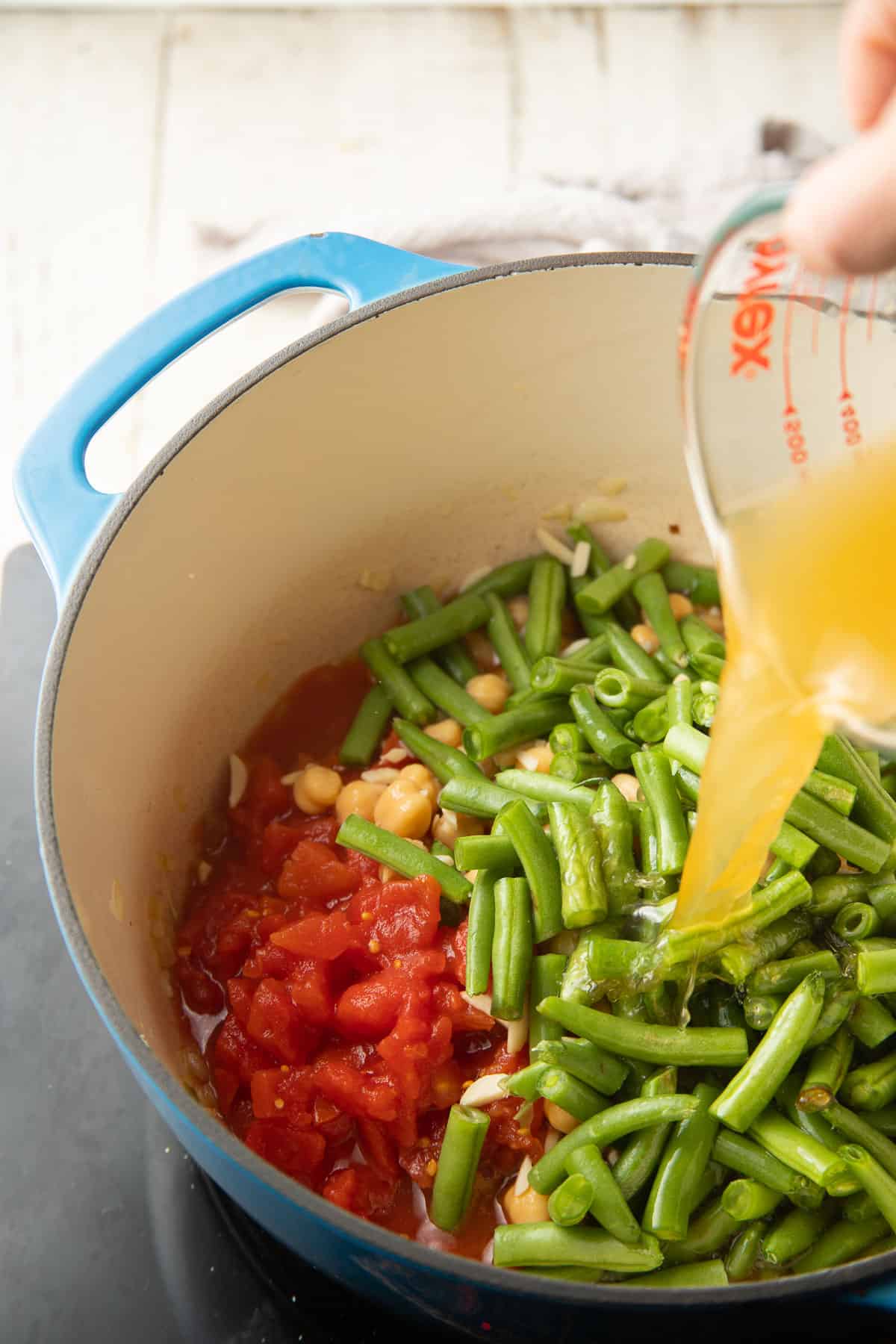 Hand pouring broth into a pot of green beans, chickpeas and tomatoes.